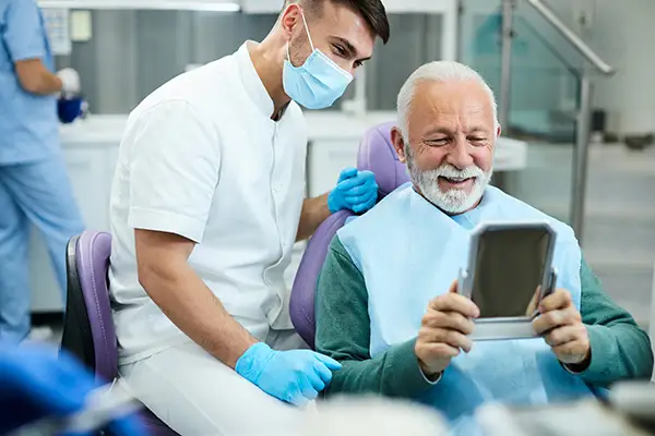 Happy, senior patient looking at himself in a mirror with a pleased, male dental assistant